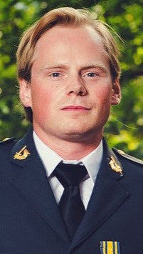 Man in a military uniform standing outdoors with greenery in the background